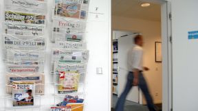 Zeitungsständer an der Wand neben Tür, durch die man Flur einen laufenden Mann sieht